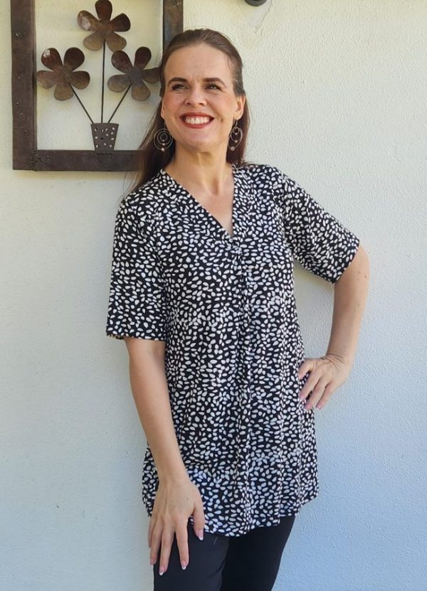 Woman smiling while wearing a stylish Black and White Viscose Lycra Front Pleat Top, featuring a comfortable and flattering fit, paired with black pants. The lightweight fabric showcases a seed-like pattern, perfect for a casual yet polished look.