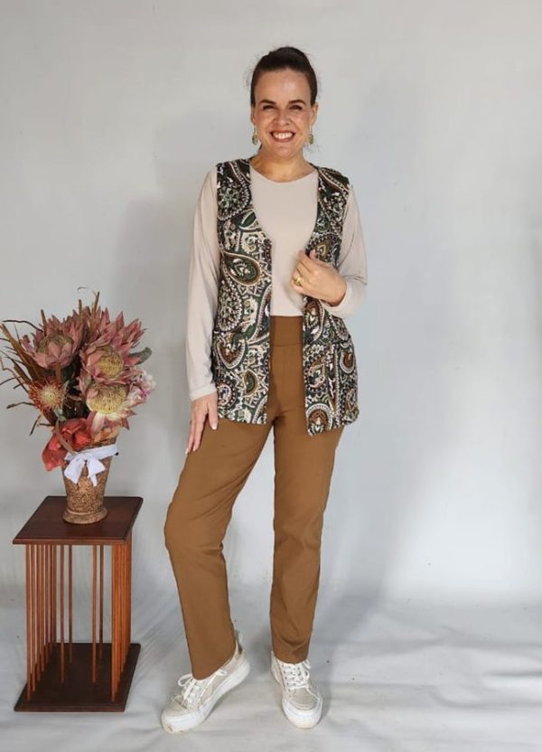 Woman wearing a J&S Creations outfit featuring the Nina Cardigan with buttons (sleeveless) in a rich paisley print, layered over the Anja Basic Top in soft cream Bon-Bon fabric, paired with Bengaline Relaxed Straight Pants in caramel. She’s wearing white sneakers and smiling brightly against a neutral backdrop, with a small wooden table and a protea flower arrangement beside her.