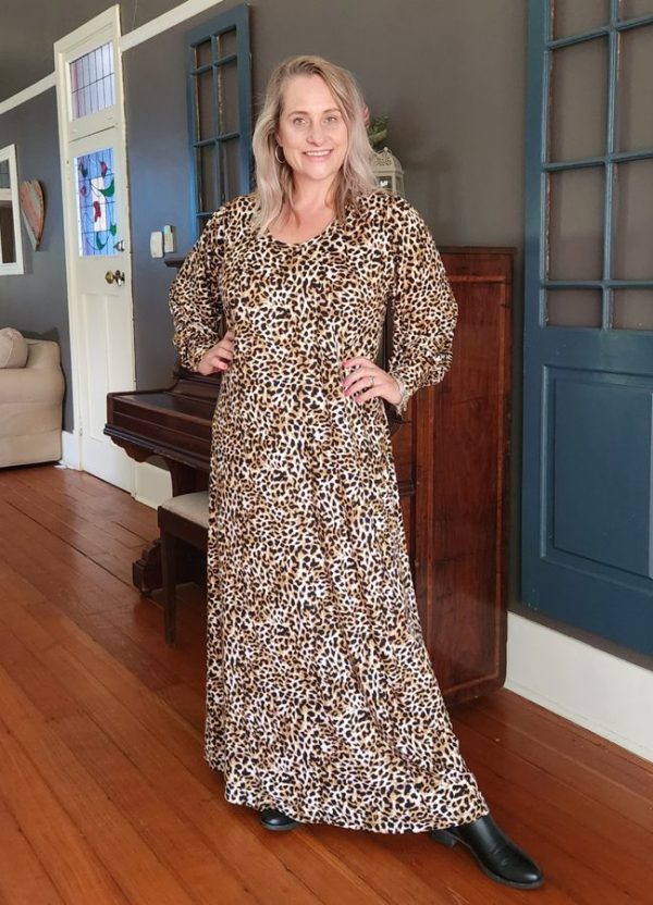 Smiling woman wearing a custom-made full-length leopard print Anja Maxi Dress by J&S Creations, styled with long puff cuff sleeves in soft DTY brush knit fabric. Photographed indoors on wooden flooring, showcasing flattering, comfortable fashion for South African women. Available in sizes Small to 6XL.