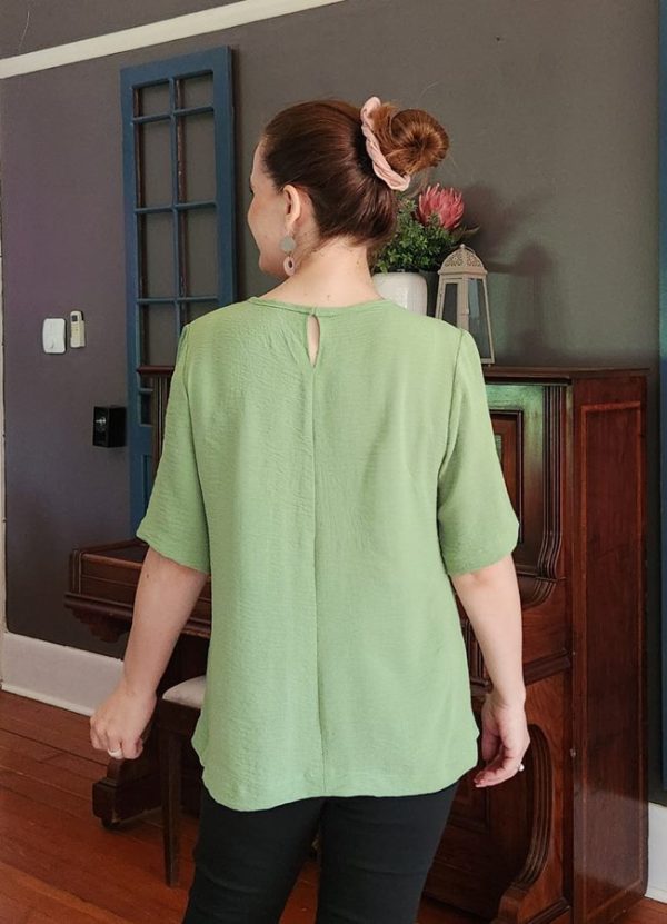 A smiling woman wearing the Elzani Airflow Pleat Top in soft olive, showcasing its breathable fabric, box pleat detail, and half sleeves. Perfect for South African women seeking comfortable, handmade fashion by J&S Creations Clothing.