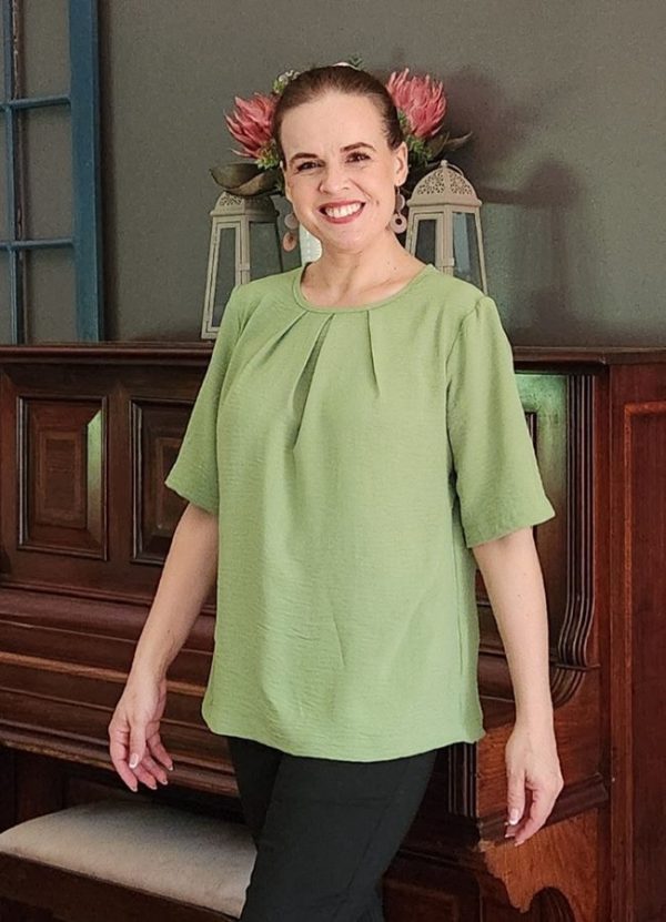 A smiling woman wearing the Elzani Airflow Pleat Top in soft olive, showcasing its breathable fabric, box pleat detail, and half sleeves. Perfect for South African women seeking comfortable, handmade fashion by J&S Creations Clothing.