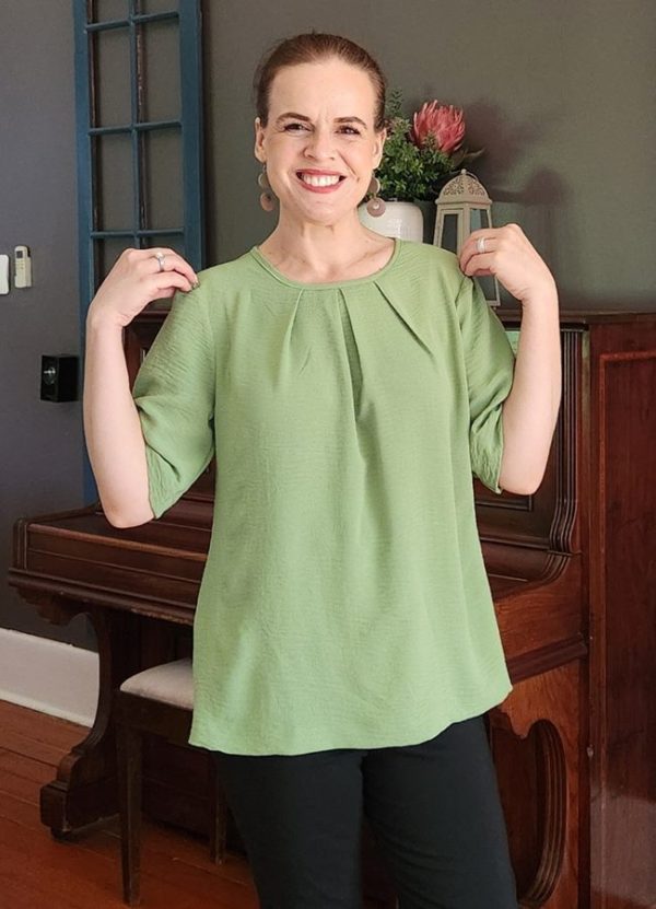 A smiling woman wearing the Elzani Airflow Pleat Top in soft olive, showcasing its breathable fabric, box pleat detail, and half sleeves. Perfect for South African women seeking comfortable, handmade fashion by J&S Creations Clothing.