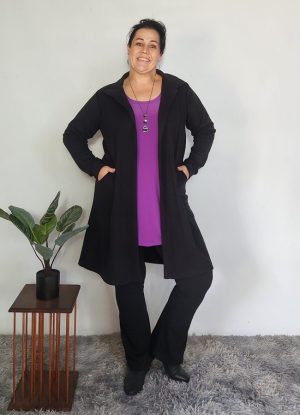Woman wearing the Zelda Cosy Collar Pocket Cardigan in black Cosy Spandex knit. The cardigan features a soft collar and a relaxed fit, styled over a bright purple top and paired with black pants and black boots. She stands confidently on a grey rug next to a small wooden plant stand with a potted plant.